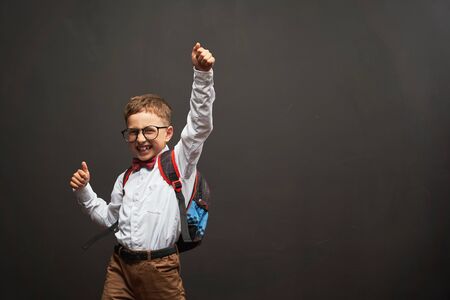 joyful little boy, on a black background with a briefcase behind his shoulders shows a gesture of victory, joy of success. Happy emotional child pupil rejoices back to school. free text, copy spaceの写真素材