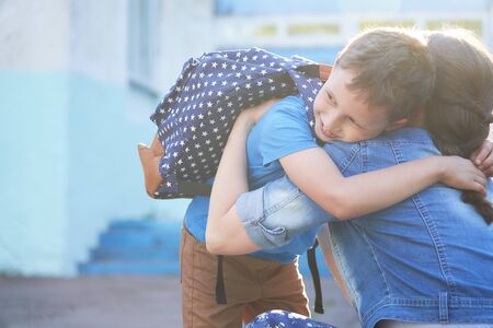 Back to school. Happy mother and son embrace in front of the elementary school. The parent takes the child to primary school. The student goes to study with a backpack. The first day of autumn.の写真素材