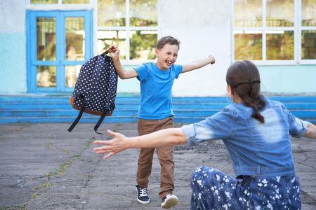 mom meets her son from elementary school. joyful child runs into the arms of his mother. a happy schoolboy runs towards his mother holding a school bag in his hands. the end of the school dayの写真素材