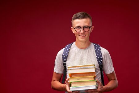 portrait of a boy student with a backpack and a stack of books in his hands smiling on a red background. funny positive teenager is a high school student with glasses. back to school. copy spaceの写真素材