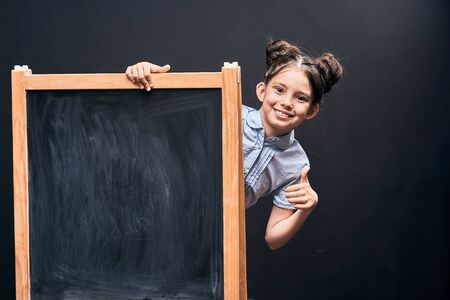the child shows a sign of approval standing at the school Board. positive schoolgirl peeking out from behind a black school Board for drawingfree text, copy space. back to schoolの写真素材