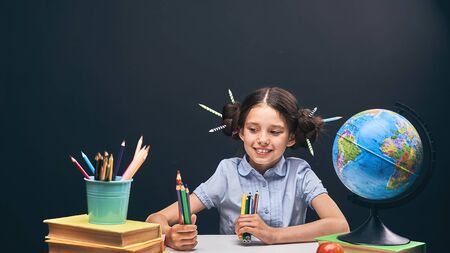 joyful little girl sitting at the table with pencils and textbooks. Happy child pupil doing homework...