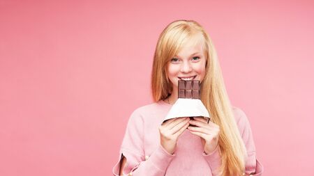 Young beautiful blonde with chocolate. teen girl bites chocolate. the temptation to eat forbidden chocolate. cheerful positive young girl holding a chocolate bar. Studio, pink background. copy spaceの写真素材