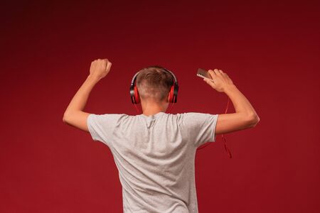 a positive teenage guy with headphones, listening to music from his smartphone on a red background, dancing with his hands up, turning his back. Rear view.の写真素材