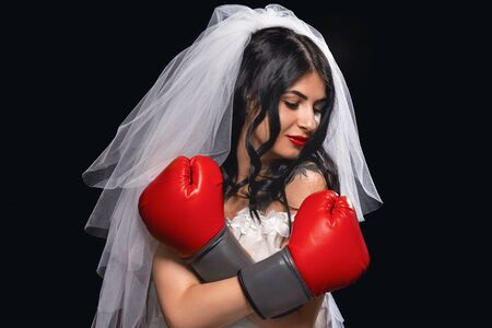 portrait of an attractive brunette with red lipstick, in a wedding dress, veil and Boxing gloves. young girl in the role of a bride on black background, arms crossed, protects herself and looks away.の写真素材