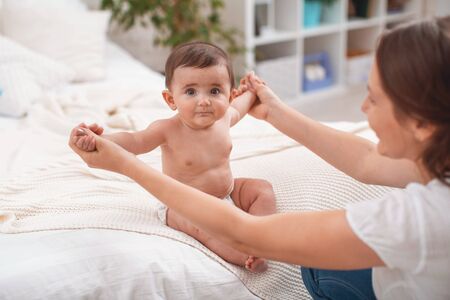 Cute baby sitting with support on bed. mother supports child sitting, spreading her hands to side. child is just starting to sit on its own. concept of development of new skills, support and care.の写真素材