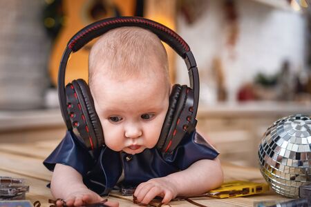 Cute little baby is lying on his tummy with headphones and listening to music. small child in leather vest with metal rivets, looking away, next to it is a mirror disco ball Blurred background, bokeh.の写真素材