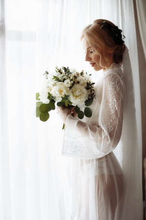 Portrait of a beautiful bride with wedding bouquet by the window. Blonde girl with curly hair and fashion makeup. High quality photoの写真素材
