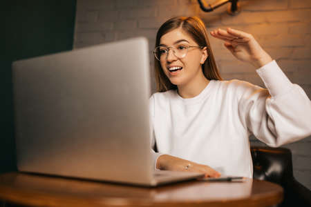 Happy young woman on a video call via a laptop computer sitting at a table in a restaurant waving...