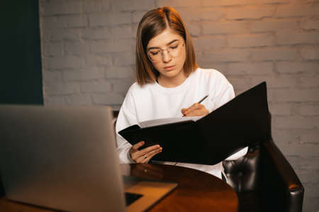 young woman with glasses communicates and write down information through laptop video communication,...