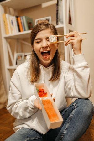 A beautiful young girl in a white sweater holds Chinese rolls and sushi with chopsticks near her eyes. High quality photoの写真素材