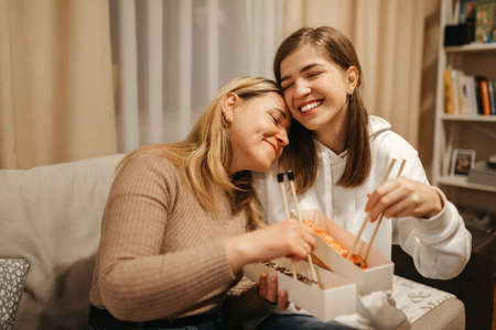 Beautiful girls friends are sitting on the couch at home and eating sushi rolls. High quality photoの写真素材