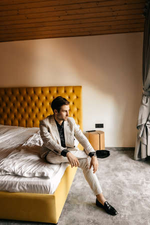 Handsome respectable groom is sitting on the bed of a luxury hotel room, the groom's light wedding suit. High quality photoの写真素材