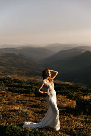 Beautiful bride in a white wedding dress silhouetted from the rays of the sun, on a background of mountainsの写真素材