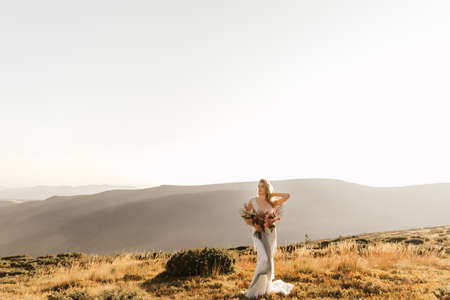 Spring wedding in the mountains. A young girl in a white dress stands on the mountain and holds a bouquetの写真素材