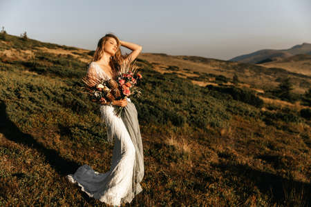 Beautiful bride in a white wedding dress with a bouquet in her hands on a background of mountainsの写真素材