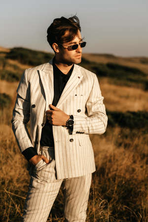 Handsome man in glasses and a light beige suit stands posing against a background of mountains. High...