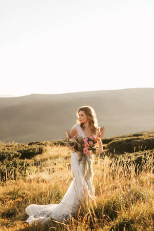 Beautiful bride in a white wedding dress stands posing and smiles in the mountains with a bouquet. High quality photoの写真素材