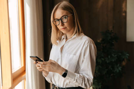 A beautiful girl in glasses stands by the window in the office in her hands with a phone typing text. High quality photoの写真素材
