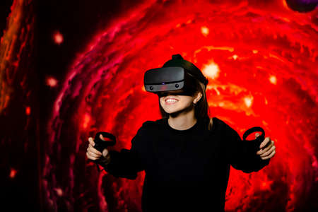 A young girl plays a game in virtual reality glasses holding gamepads in her hands. Bright red background in the playroom. High quality photoの写真素材