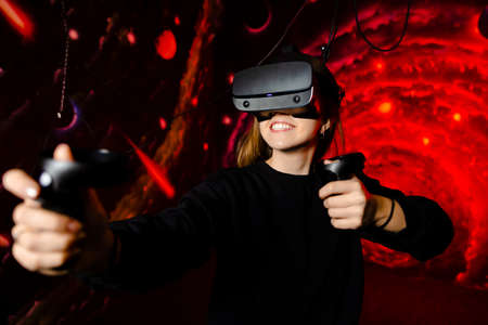 Young beautiful girl in reality reality glasses, glasses for video games, holding gamepads in hands. On a red starry space background. High quality photoの写真素材
