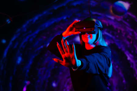 A young girl using a virtual reality helmet plays. Neon red and blue lights on a space background. Holding hand outstretched forward, stop sign.の写真素材