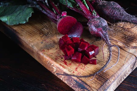 three juicy beets with leaves, one of which sliced lies next to diced beets on a wooden board....
