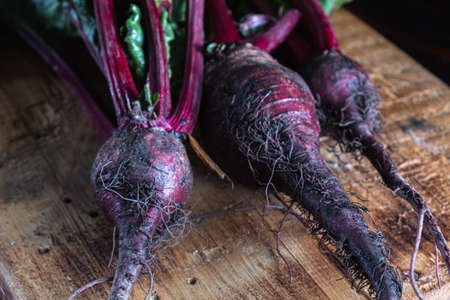three unwashed juicy beets with green leaves lies on a wooden board. country style. rusty style. Horizontal orientation. ugly food. dark background.の写真素材