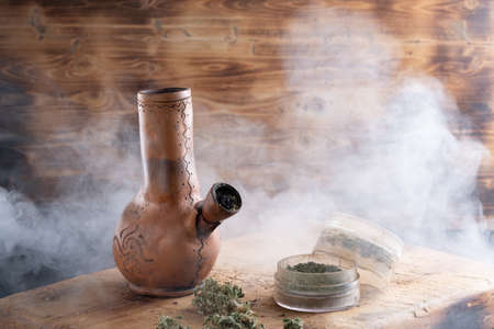 Old bong and open cannabis grinder with chopped weed and marijuana in smoke are located on the...