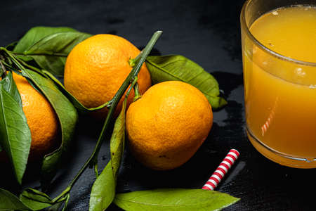 on a dark surface are three tangerines with leaves next to a glass of juice. Nearby lies a red and white striped straw.の写真素材