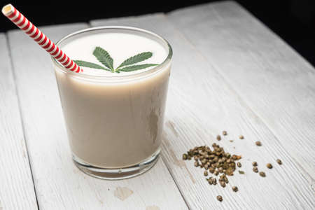 Hemp milk is poured into a glass. On the surface is a cannabis leaf. Nearby marijuana seeds. On the white old weathered boards. In a glass, red and white non-plastic straw.の写真素材