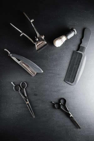 On a black dusty surface are old barber tools. Vintage manual hair clipper comb razor shaving brush shaving brush hairdressing scissors. black monochrome. vertical. top view. flat lay.の写真素材