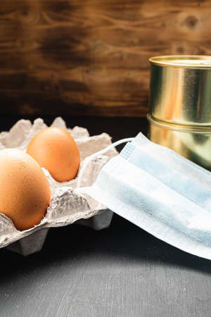 medical mask and two cans of canned food and two eggs in a package on a wooden background. vertical