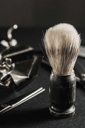 On a black surface are old barber tools. shaving brush vintage manual hair clipper, comb, razor, hairdressing scissors. black monochrome. Close-up. Barbershop background. contrast shadows. Vertical orientation.の写真素材