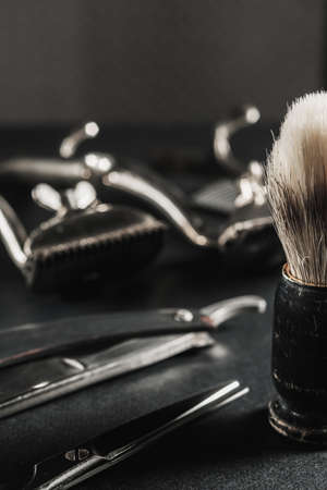On a black surface are old barber tools. shaving brush vintage manual hair clipper, comb, razor, hairdressing scissors. black monochrome. Close-up. Barbershop background. contrast shadows. Vertical orientation.の写真素材