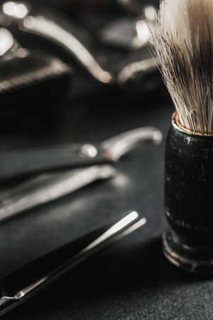 On a black surface are old barber tools. shaving brush vintage manual hair clipper, comb, razor, hairdressing scissors. black monochrome. Close-up. Barbershop background. contrast shadows. Vertical orientation.の写真素材