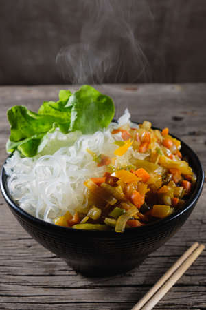 A plate of rice noodles stands on an old wooden table top. The side dish consists of fried onions, carrots, paprika. Verticalの写真素材