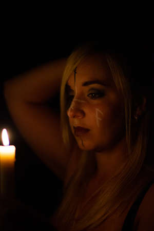 A beautiful blonde woman stands at night with a candle in her hand and looks at the flame. A witch with symbols on her face looks thoughtfully at the candle flame. Halloween conceptの写真素材