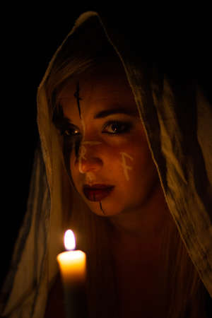 A woman in the Gothic image of a witch in a white hood in the dark holds a burning candle in front of her. Woman witch with magical patterns on her face looks at the camera, prepares for halloweenの写真素材