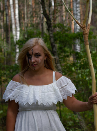 The wind blows the blond hair of a witch in the forest preparing for Halloween. A witch in a white dress with a staff in her hand with makeup on her face looks defiantly into the camera. Halloweenの写真素材