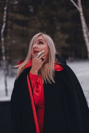A halloween woman stands in a winter forest in a black cloak and hood and looks up thoughtfully. Witch in a red dress in the forest with rings on her hands. Halloween conceptの写真素材