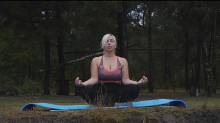 With her legs and arms folded in the lotus position, a beautiful woman meditates in nature. A young blonde woman practices yoga in the early morning at the edge of the forest.の写真素材