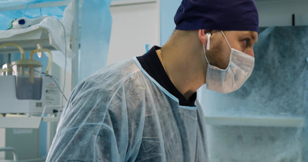 The operating surgeon listens to music through a wireless earpiece during the operation. A male doctor works in an operating room in a surgical mask and cap.の写真素材