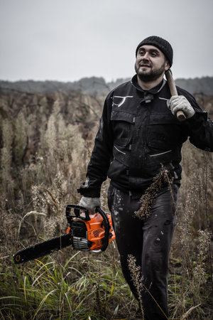 A bearded man carries a chainsaw and an ax in his hands. Having finished sawing the trunks of fallen trees, the men gathered the tool and went to the car. Front view.の写真素材