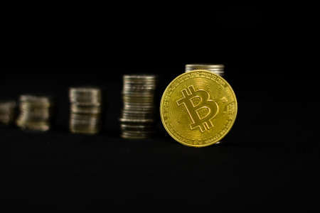 A bitcoin coin stands leaning on a stack of coins on a black isolated background. Golden bitcoin...