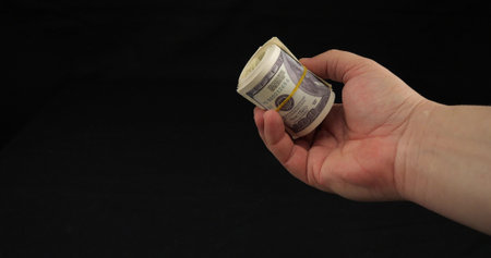 On a black background, a hand holds a roll of hundred dollar bills. Place for insertion, close-up. A mans hand holds out a large stack of one hundred dollar bills.の写真素材
