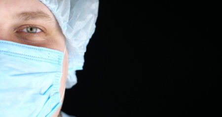 Half face of a surgeon on a black isolated background smiling cutely at the camera. A doctor in a medical mask and a surgical cap smiles at the camera.の写真素材