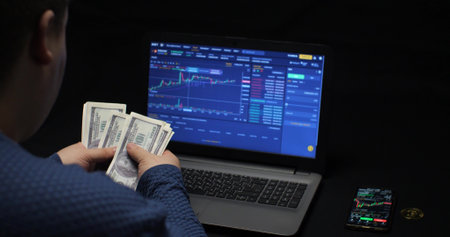 A trader with dollars in his hands against the background of a cryptocurrency chart on a laptop screen. Earnings from trading on a cryptocurrency exchange are held by a trader.の写真素材