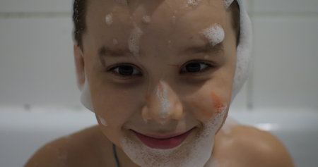 Child boy smiling while looking at camera in foam. Close-up of the childs face in the foam in the bathtub. The child enjoys bathing in a bubble bath. Concept of childrens emotions.の写真素材