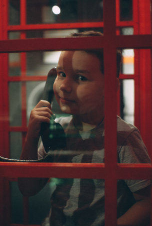 Child boy in a red phone booth smiles and looks at the camera through the window. The boy holds in his hand the receiver of an old telephone in a red traditional telephone booth.の写真素材